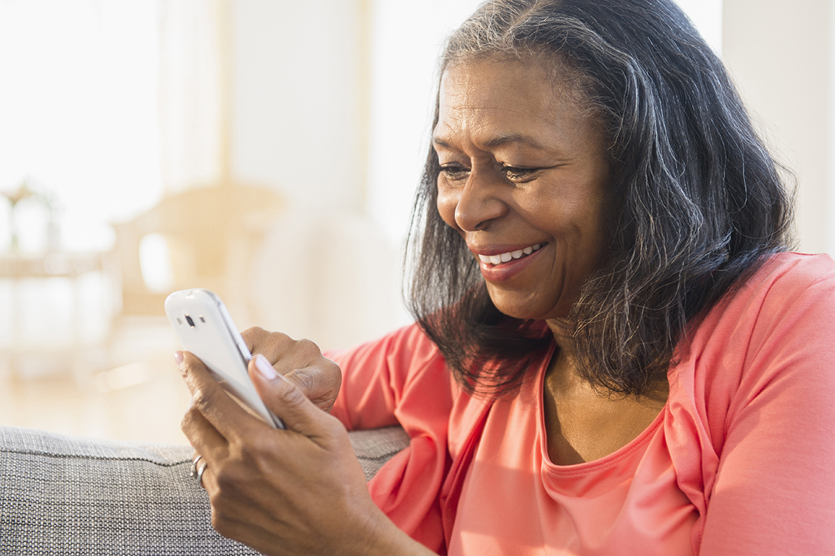 A smiling women sitting on her couch with her arm over the back and using her smart phone