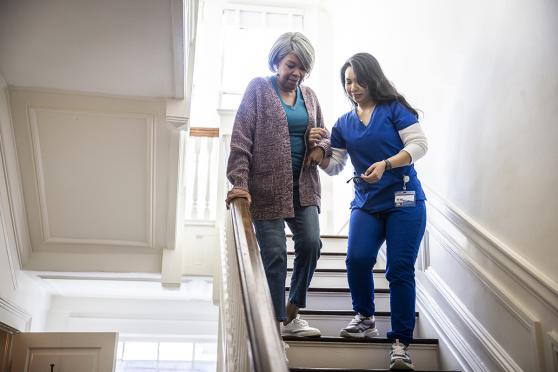 A home health aide helps a senior walk down the stairs