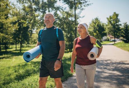 Two older people walk through a park with yoga mats