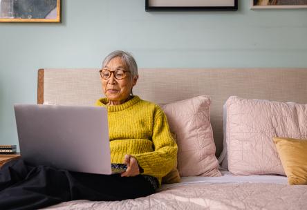 A senior sits on a bed with a laptop