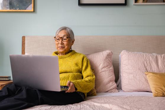 A senior sits on a bed with a laptop